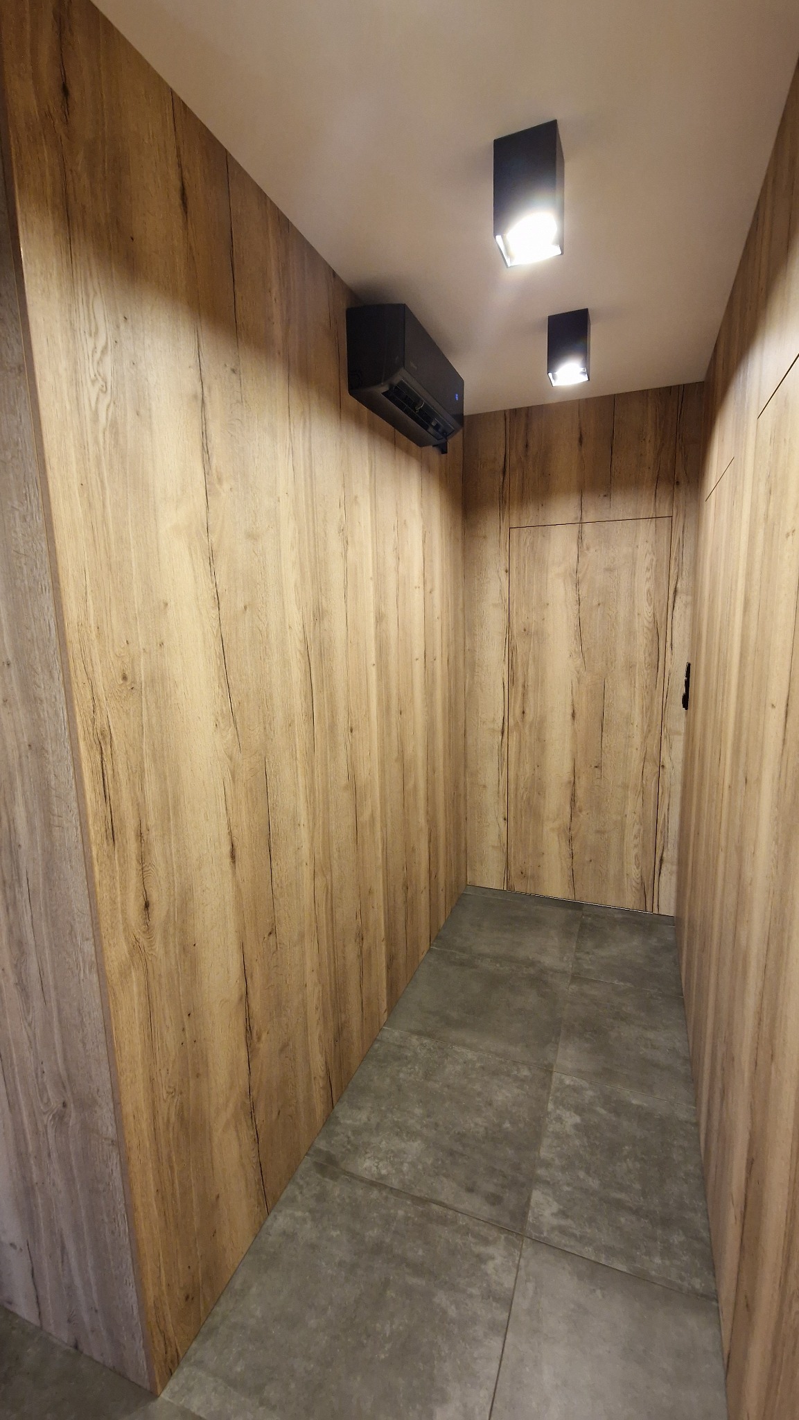Wnętrze korytarza wykończonego drewnopodobnymi panelami ściennymi, z zamontowanym czarnym klimatyzatorem sufitowym i dwoma czarnymi reflektorami punktowymi, szara podłoga z płytek.
