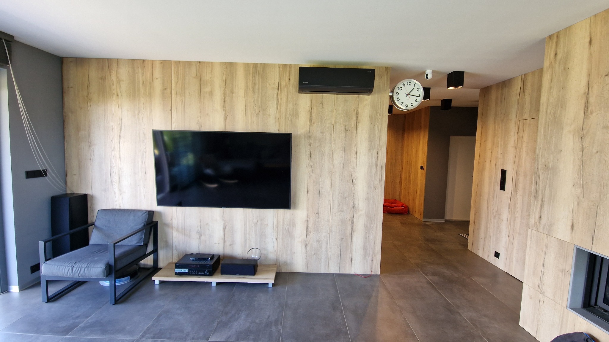 Nowoczesny salon z klimatyzatorem ściennym w kolorze czarnym, zamontowanym nad telewizorem na ścianie z drewnianą okładziną.