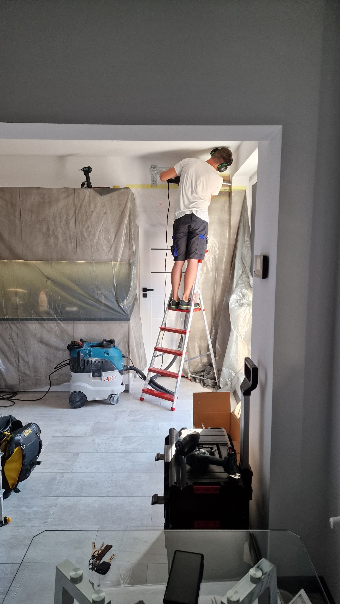 Instalator w słuchawkach na drabinie wierci otwór w suficie pod instalację, pomieszczenie zabezpieczone folią malarską, widoczny odkurzacz przemysłowy i skrzynka z narzędziami.