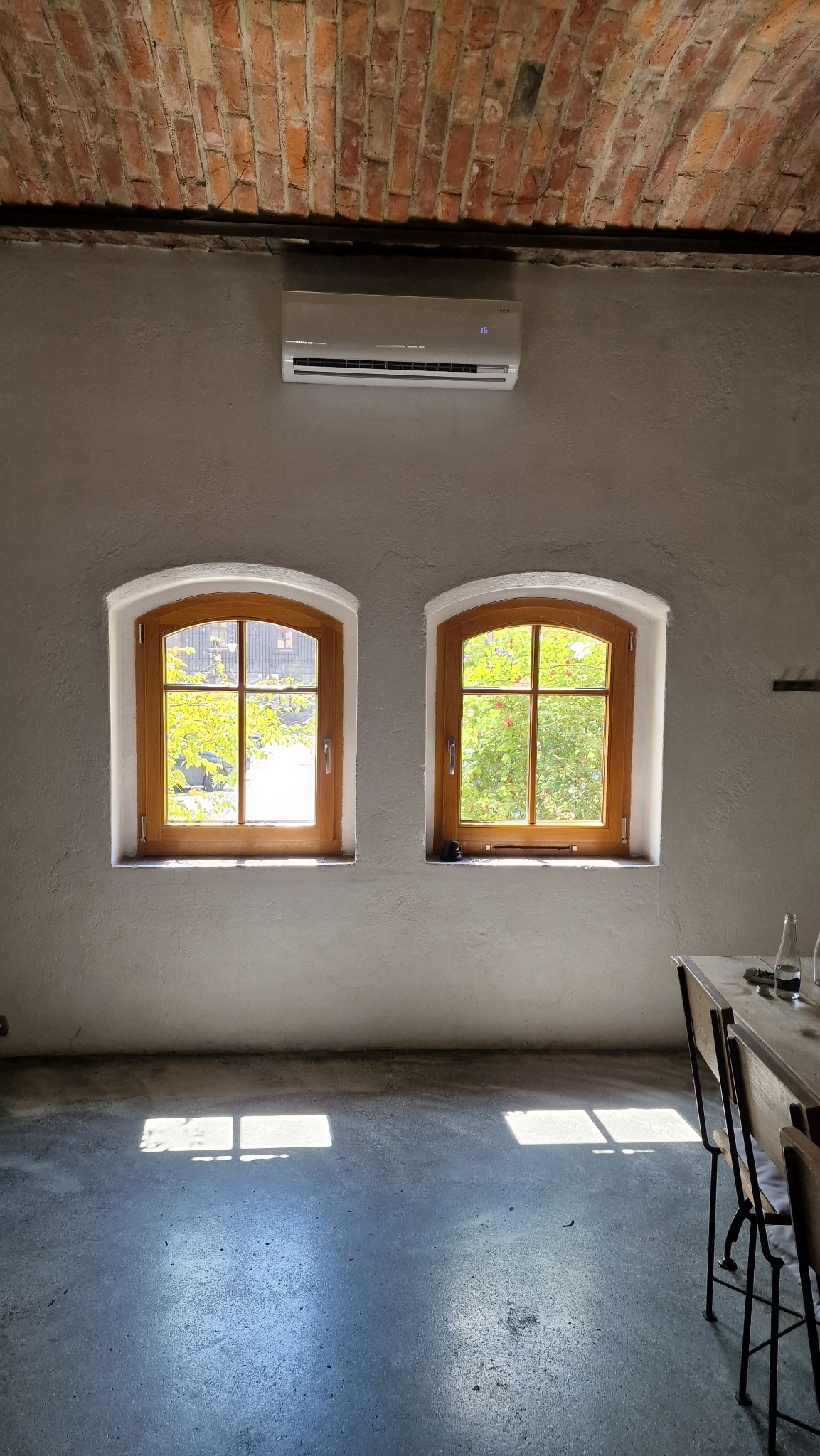 Wnętrze z zamontowanym klimatyzatorem ściennym nad dwoma drewnianymi oknami łukowymi, ceglany sufit, betonowa podłoga z refleksami światła słonecznego.