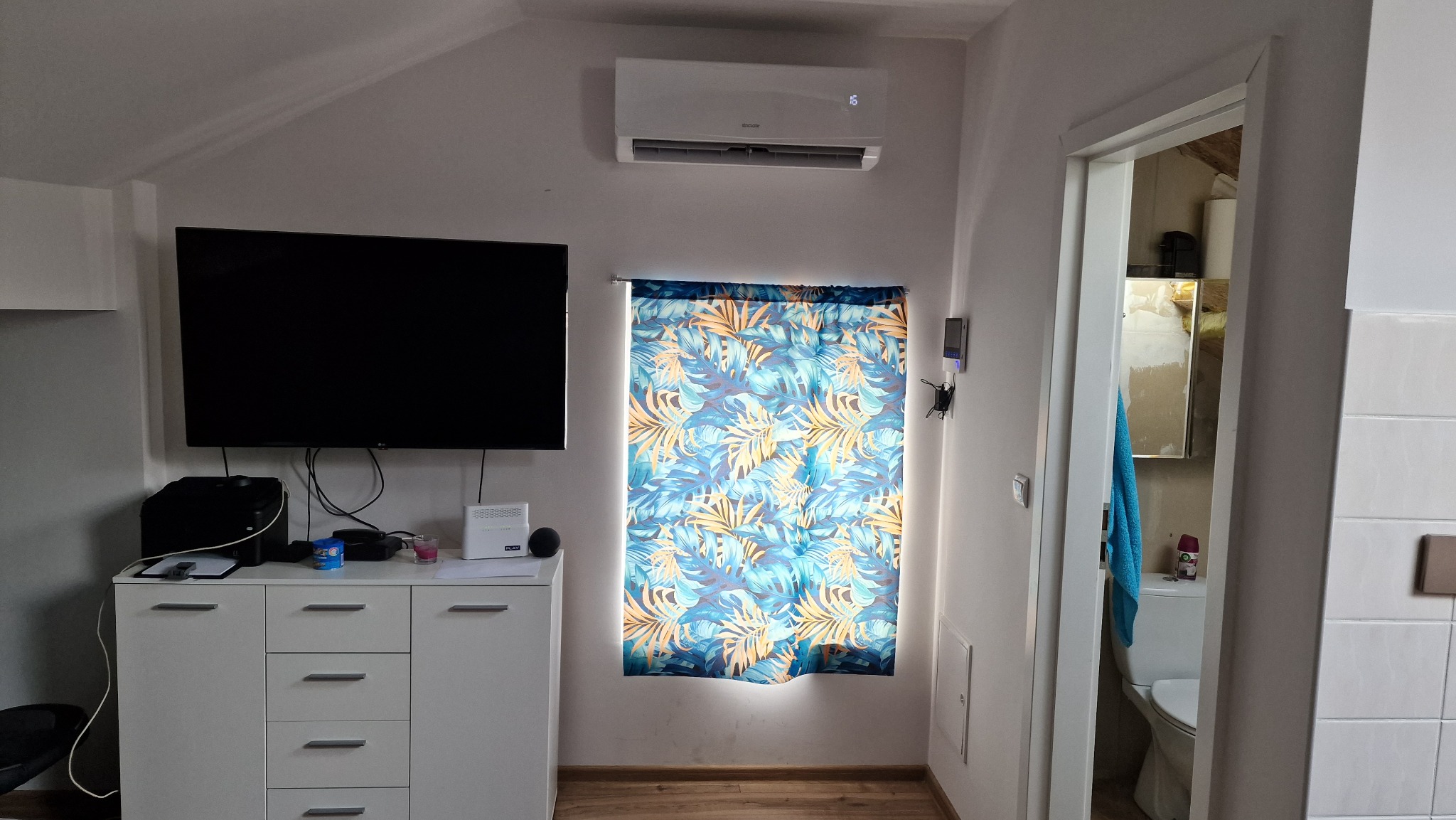 Wnętrze pokoju na poddaszu w Siemianowicach Śląskich z zamontowaną klimatyzacją na ścianie, nad oknem z zasłoną w tropikalne wzory. Obok okna widoczny telewizor i biała komoda.