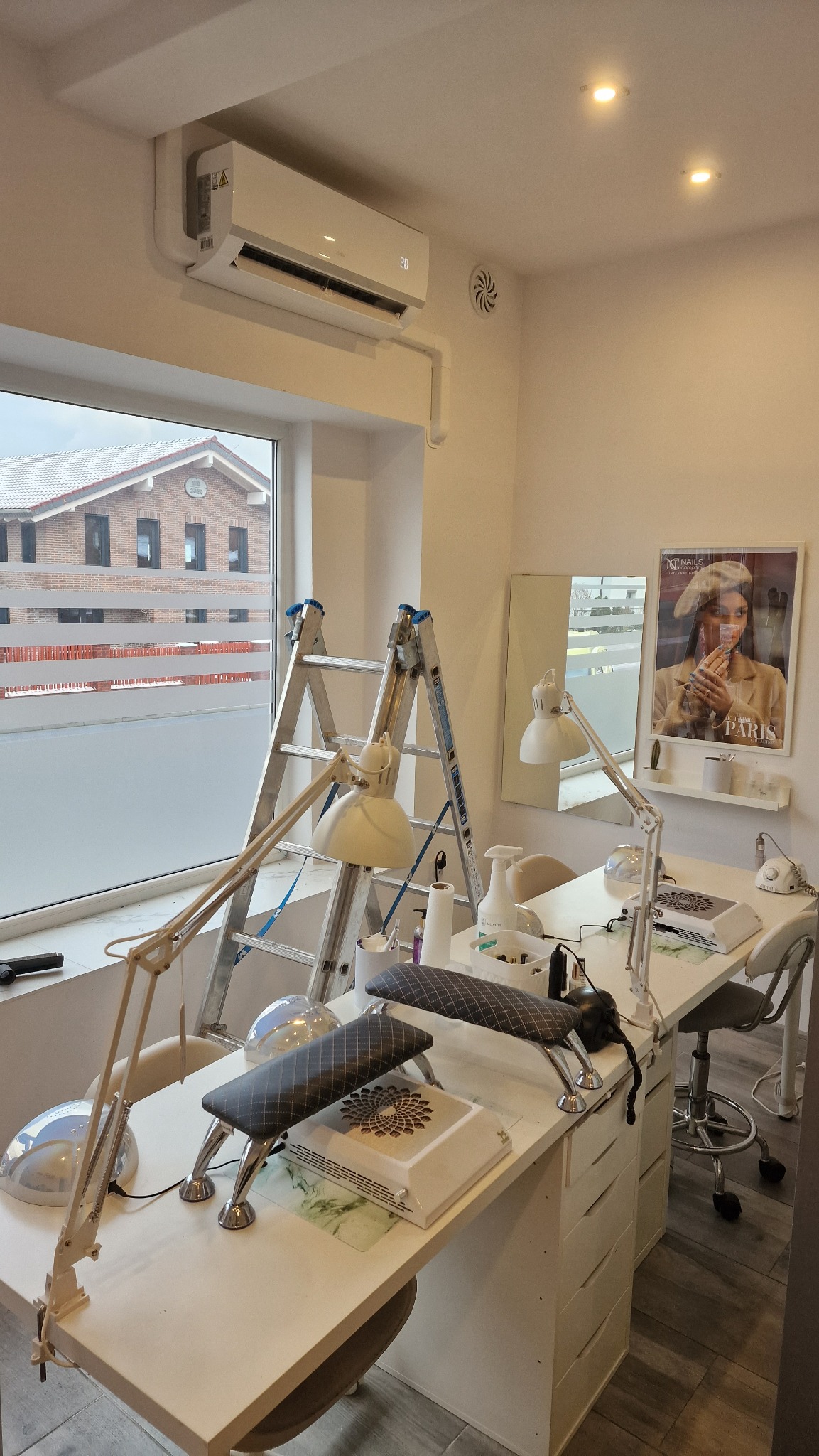 Biały klimatyzator zamontowany nad oknem w salonie kosmetycznym, widoczna drabina i stanowiska do manicure.