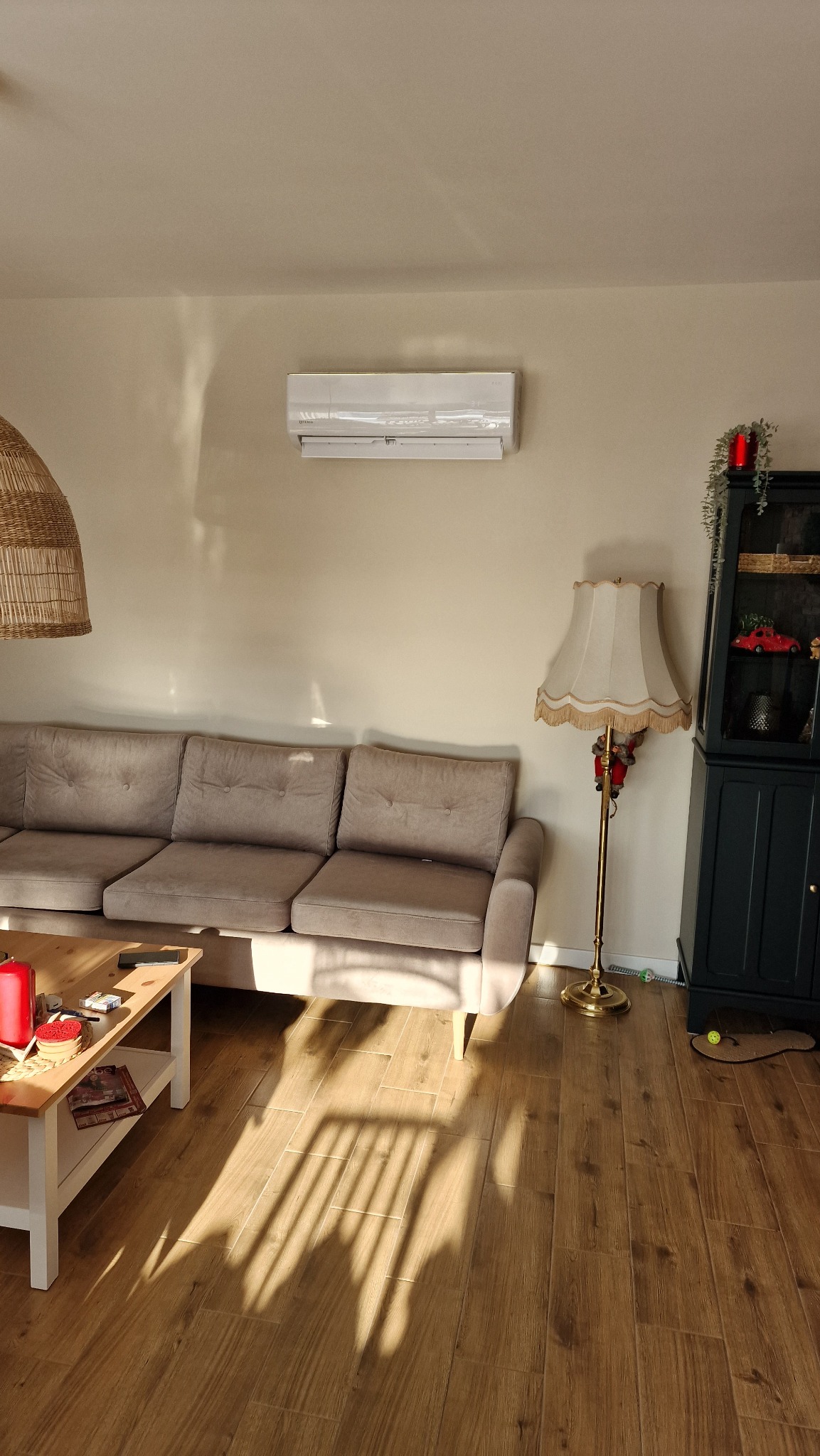 Biały klimatyzator ścienny zamontowany w salonie nad beżową sofą i drewnianą podłogą z odbijającym się światłem słonecznym.