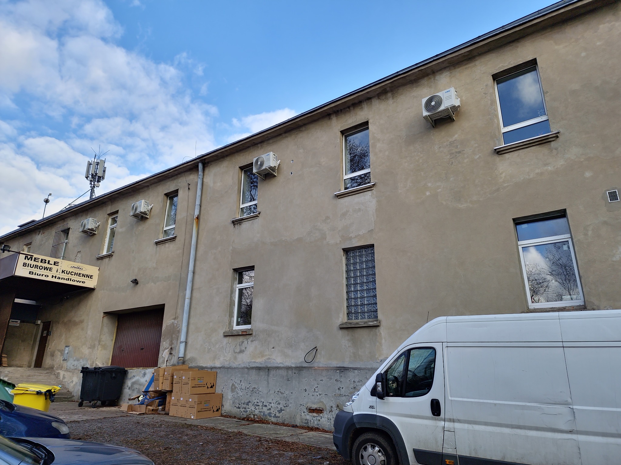 Zewnętrzny widok budynku biurowego z zamontowanymi klimatyzatorami na ścianie, widoczny biały samochód dostawczy oraz anteny na dachu.