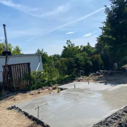 Świeżo wylany fundament z wystającymi rurami instalacyjnymi, widoczne szalunki i otaczający teren przygotowany pod dalsze prace budowlane, w tle zieleń drzew i budynek gospodarczy.