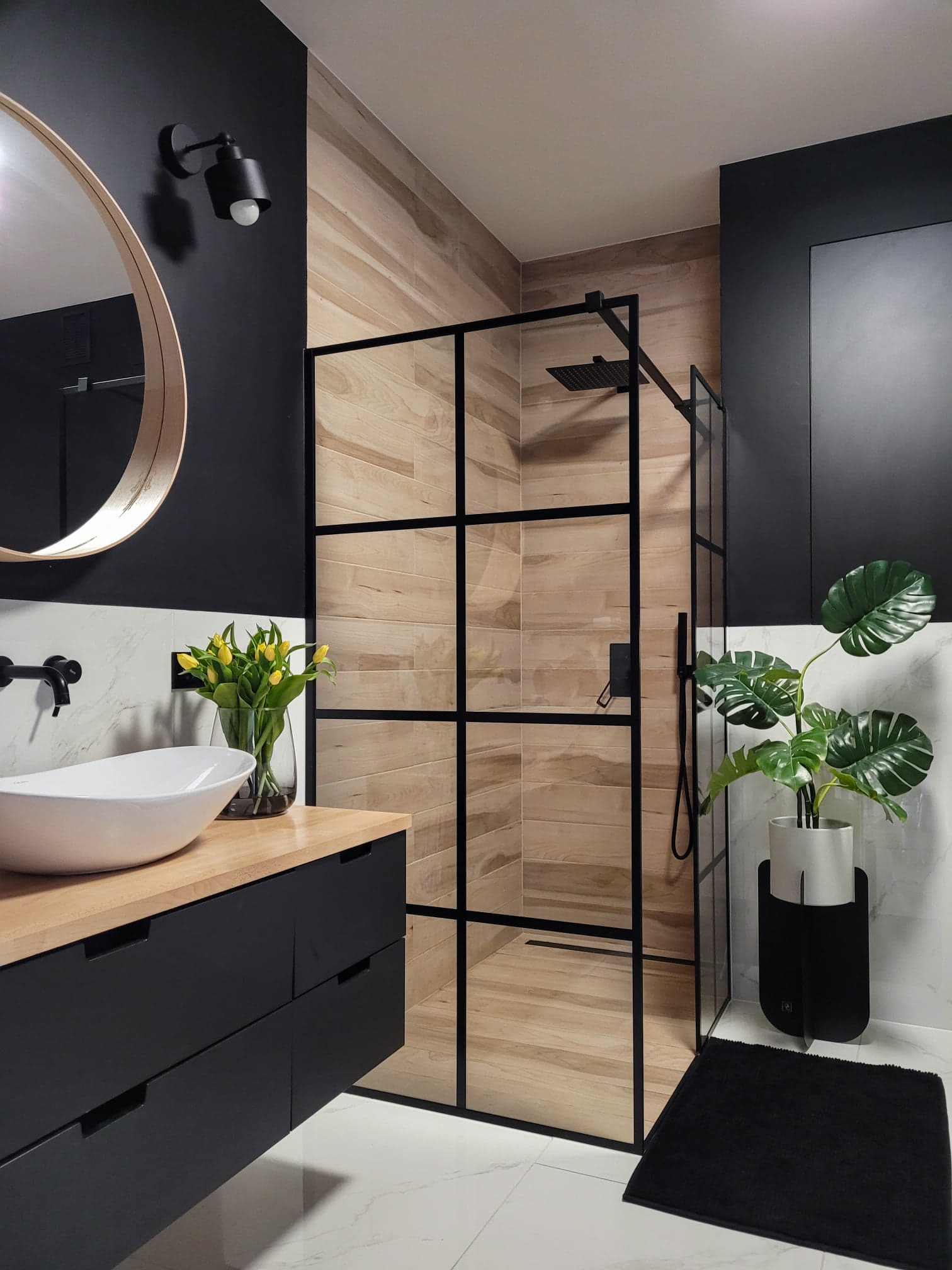 Nowoczesna łazienka z kabiną prysznicową typu walk-in z czarnymi szprosami, umywalką nablatową na drewnianym blacie i czarnymi dodatkami, uzupełniona o żółte tulipany w wazonie i zieloną roślinę...