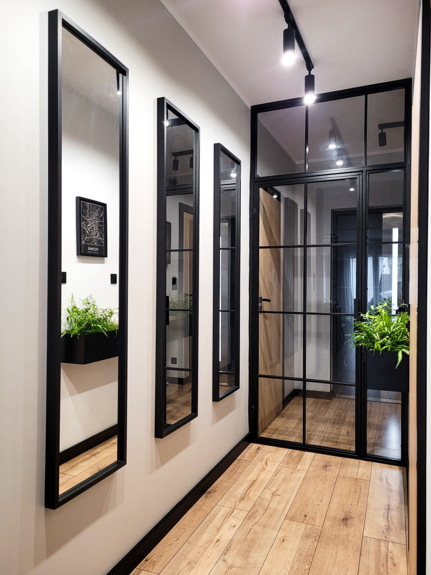 Nowoczesny korytarz w Tarnowie Podgórnym z trzema lustrami w czarnych ramach, rośliną w wiszącej donicy i przeszklonymi drzwiami w stylu industrialnym.