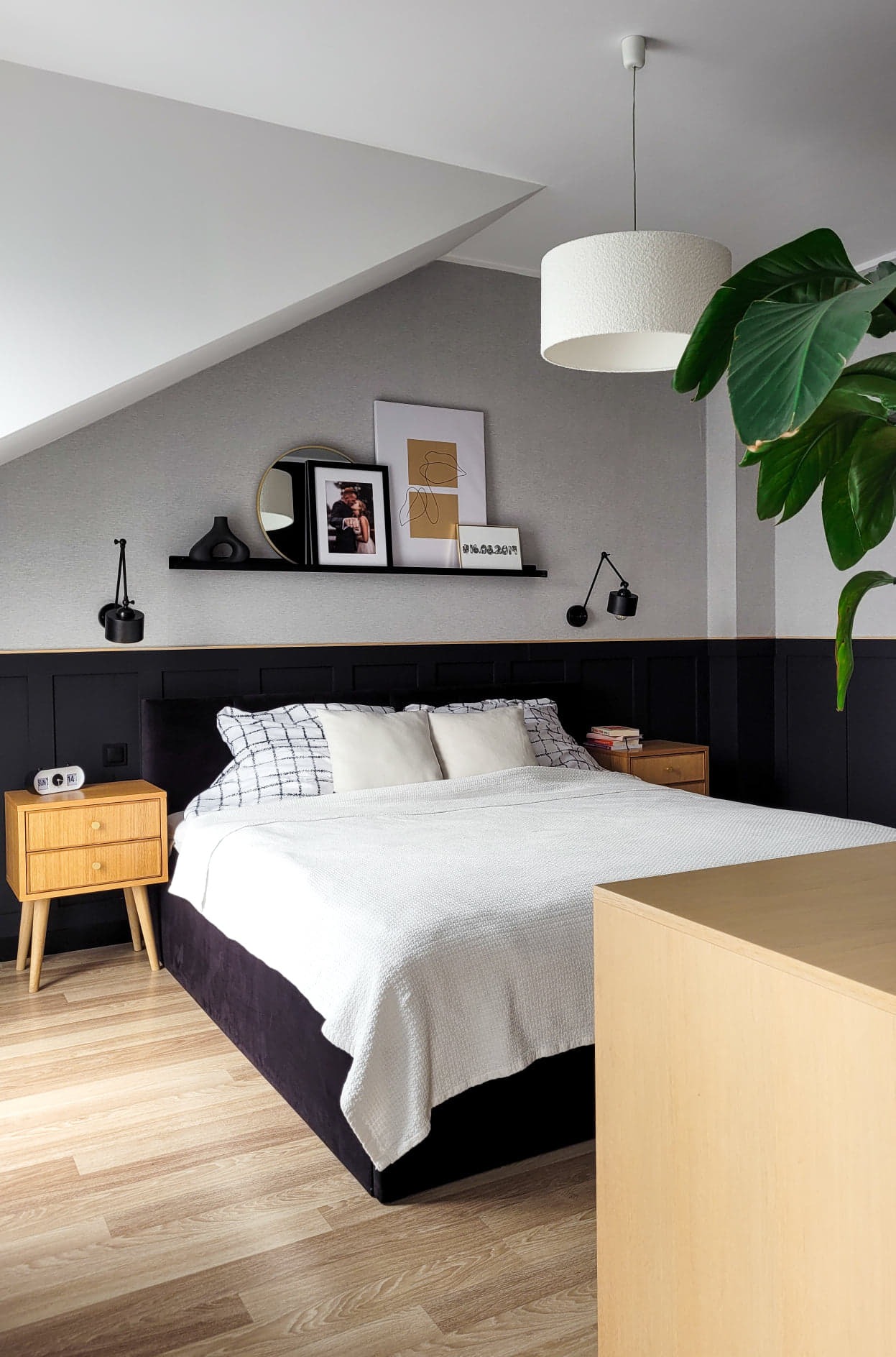 Sypialnia na poddaszu w stylu skandynawskim: łóżko z czarną ramą, biała pościel, drewniane szafki nocne, czarna półka z dekoracjami, lampy ścienne, roślina doniczkowa, oraz skośny sufit.