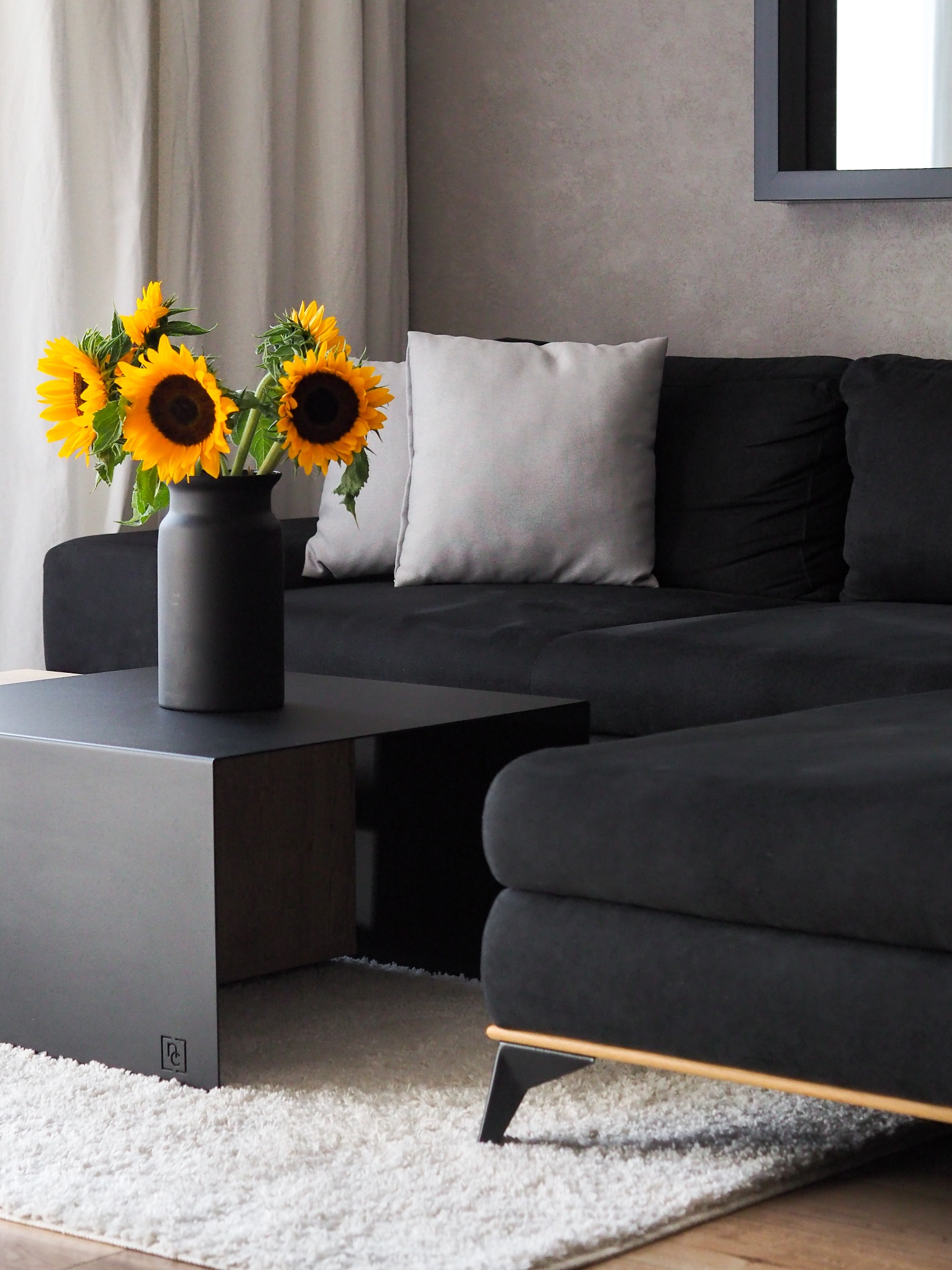 Nowoczesny salon: czarna sofa narożna z jasnymi poduszkami, czarny stolik kawowy z wazonem słoneczników, puszysty biały dywan.