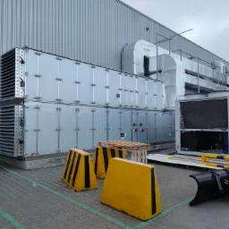 Zewnętrzna instalacja wentylacyjna z dwoma modułami rekuperacji i systemem rur, widoczne żółte bariery ochronne i pług do śniegu w tle, całość na tle szarego budynku przemysłowego.