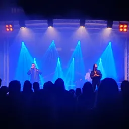 Występ duetu muzycznego na scenie oświetlonej niebieskimi reflektorami, widok zza publiczności.