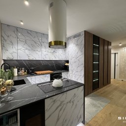 Sigmat Meble - Nowoczesna kuchnia z białymi szafkami górnymi z marmurowym wzorem, czarnym blatem i wyspą z białym marmurowym frontem, uzupełniona o drewnianą zabudowę z przeszklonymi witrynami i złotymi akcentami.