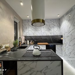 Sigmat Meble - Nowoczesna kuchnia z marmurowymi frontami szafek, czarnym blatem i białym czajnikiem na płycie indukcyjnej. Widoczny okap w kolorze białym ze złotym akcentem. Na blacie dekoracyjne złote kielichy...