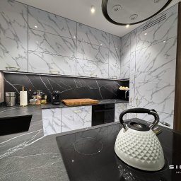 Sigmat Meble - Nowoczesna kuchnia z białymi szafkami z marmurowym wzorem i czarnym blatem, czajnik na płycie indukcyjnej, logo firmy meblarskiej w prawym dolnym rogu.