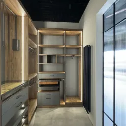 Nowoczesna garderoba z drewnianymi elementami, szarymi szufladami i oświetleniem LED, widok na czarny sufit rastrowy i grzejnik przy teksturowanej szybie okiennej.