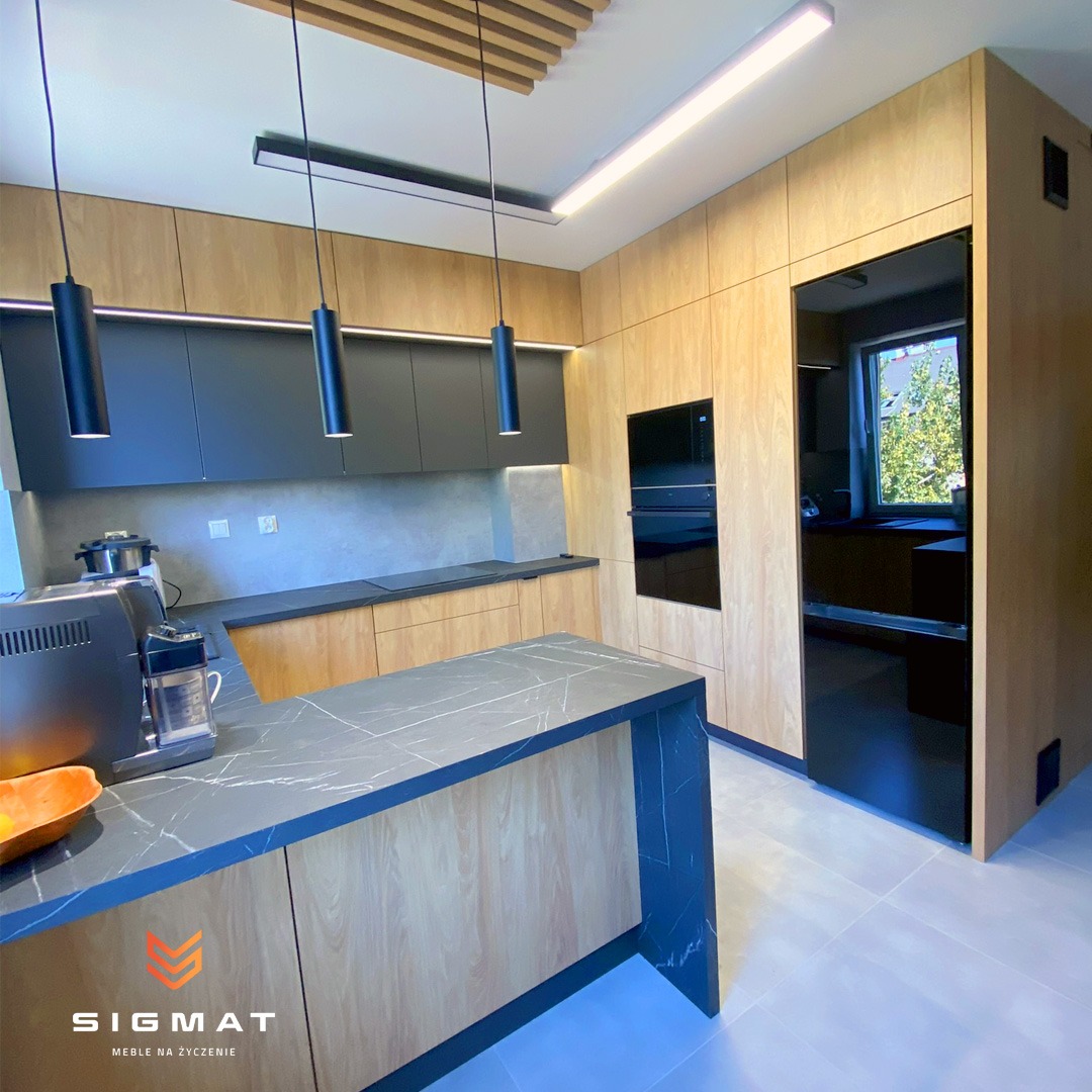 Nowoczesna kuchnia na wymiar z drewnianymi frontami i czarnymi szafkami wiszącymi, oświetlona wiszącymi lampami cylindrycznymi i listwami LED, z wyspą z blatem imitującym marmur i widocznym...