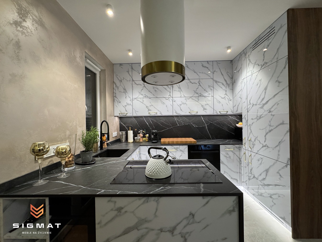 Nowoczesna kuchnia z marmurowymi frontami szafek, czarnym blatem i białym czajnikiem na płycie indukcyjnej. Widoczny okap w kolorze białym ze złotym akcentem. Na blacie dekoracyjne złote kielichy...