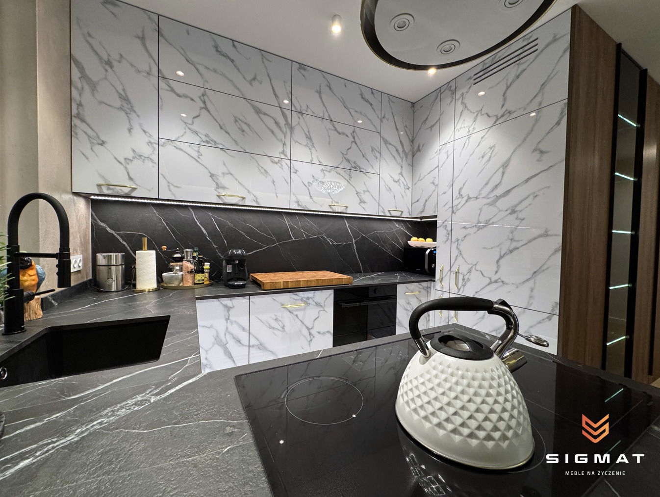 Nowoczesna kuchnia z białymi szafkami z marmurowym wzorem i czarnym blatem, czajnik na płycie indukcyjnej, logo firmy meblarskiej w prawym dolnym rogu.