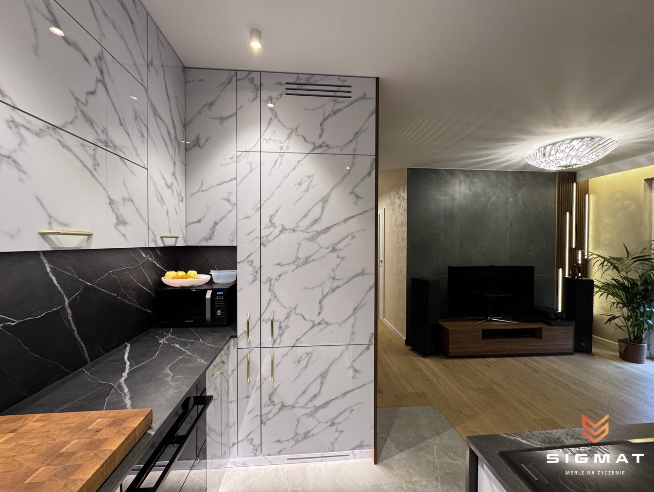 Nowoczesna kuchnia z białymi szafkami z marmurowym wzorem, czarnym blatem i złotymi uchwytami, widok na salon z telewizorem i rośliną doniczkową, oświetlenie sufitowe w obu pomieszczeniach.