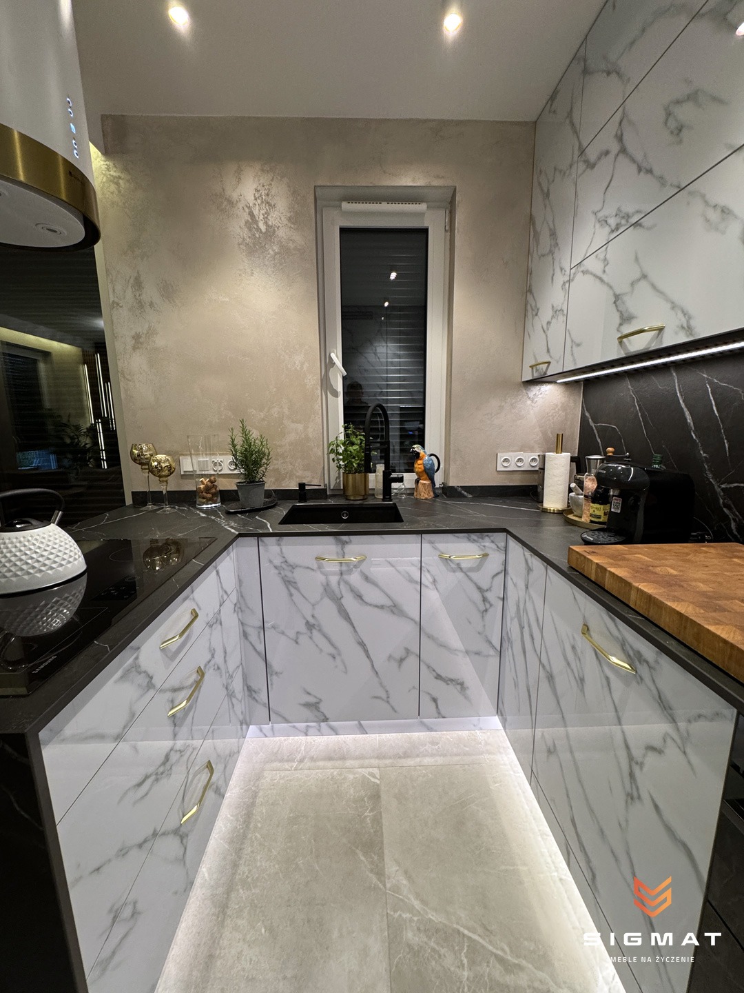 Nowoczesna kuchnia w kształcie litery U z białymi frontami szafek imitującymi marmur, złotymi uchwytami i czarnym blatem, oświetlona listwą LED pod szafkami, z widocznym oknem i elementami...