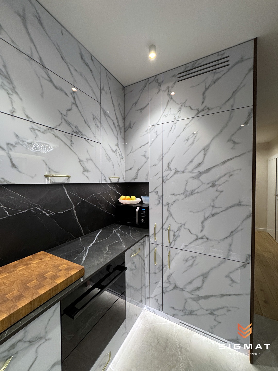 Nowoczesna kuchnia z białymi szafkami z marmurowym wzorem, czarnym blatem i złotymi uchwytami, oświetlenie punktowe i deska do krojenia na blacie.