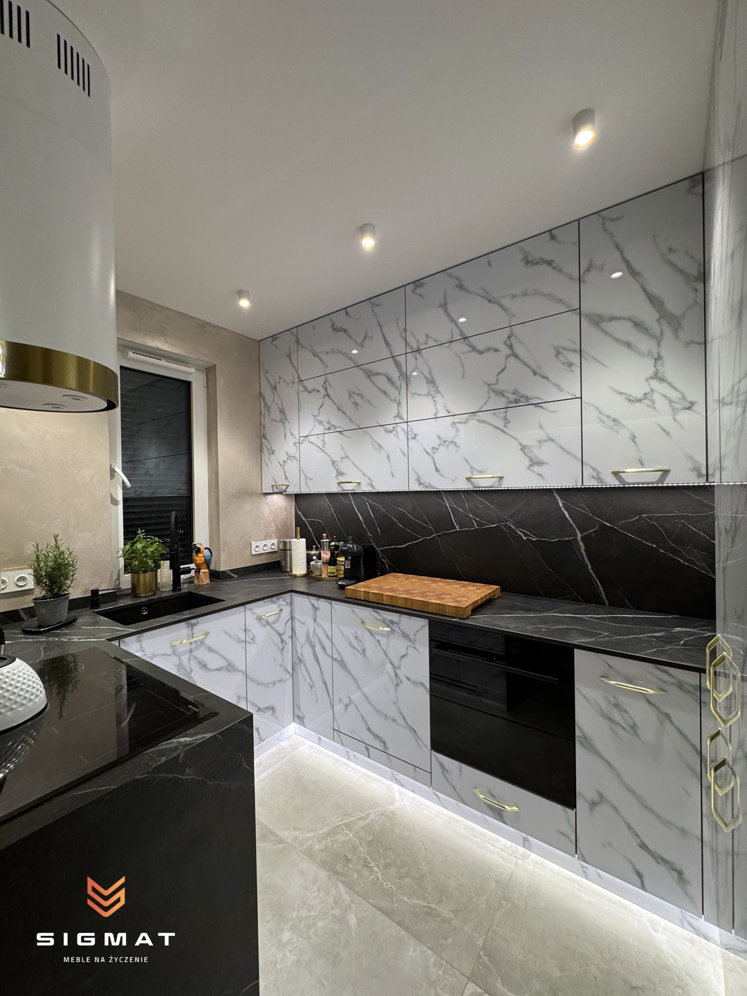 Nowoczesna kuchnia w kształcie litery U z białymi szafkami z marmurowym wzorem, czarnym blatem i złotymi uchwytami, oświetlona listwą LED pod szafkami.