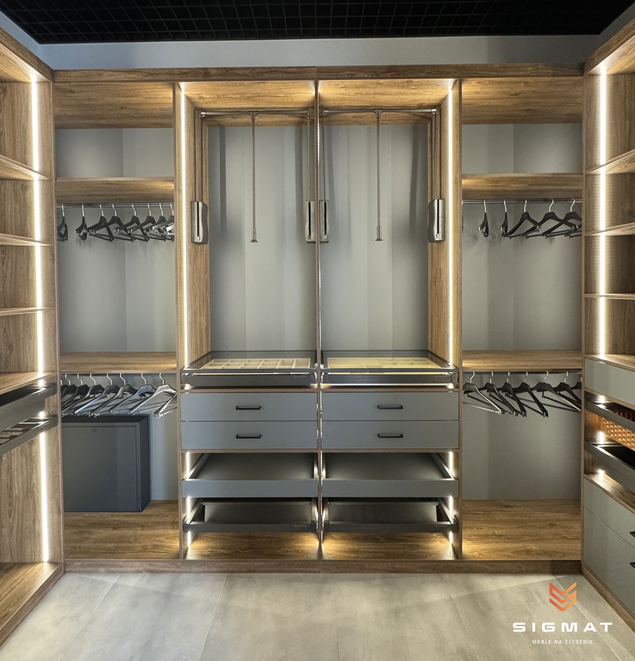 Nowoczesna garderoba z oświetleniem LED, szufladami z szarymi frontami i półkami z drewnopodobnym wykończeniem, widoczne wieszaki i sejf wewnątrz szafy.