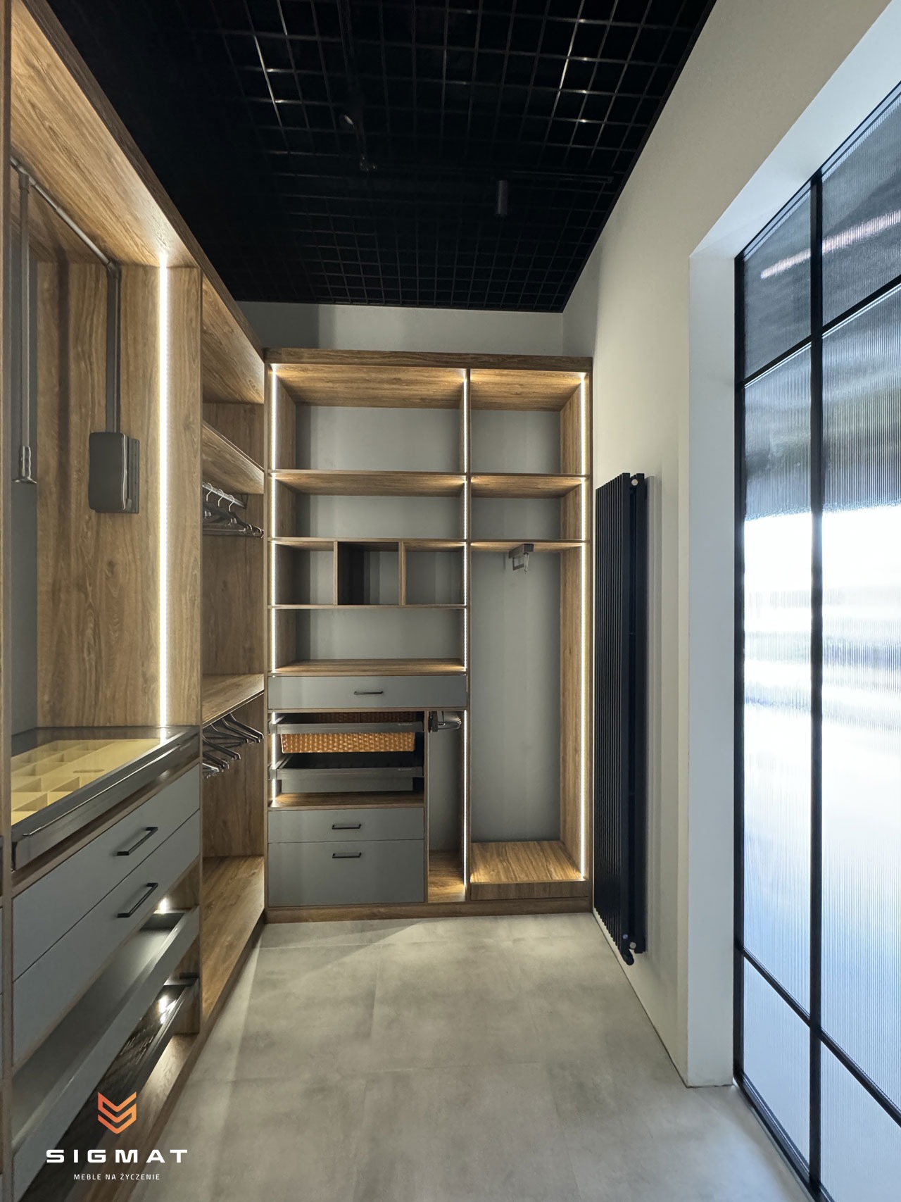 Nowoczesna garderoba z drewnianymi elementami, szarymi szufladami i oświetleniem LED, widok na czarny sufit rastrowy i grzejnik przy teksturowanej szybie okiennej.