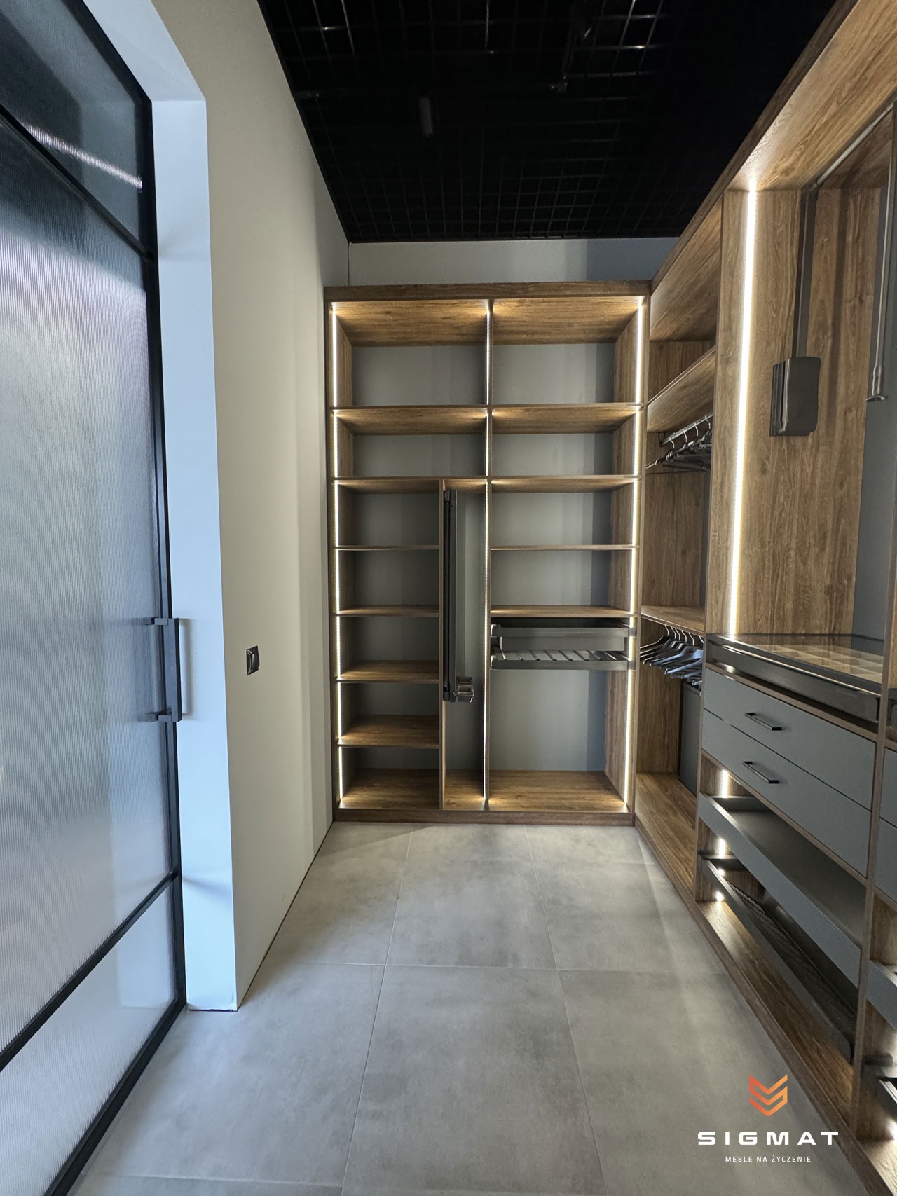Nowoczesna garderoba z podświetlanymi półkami z drewna i szarymi szufladami, widok z perspektywy wejścia przez szklane drzwi.