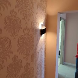 Vladyslav Vashchuk - Ściana z beżową tapetą w ornamenty i czarny kinkiet, widok na korytarz z szarymi płytkami podłogowymi.