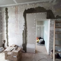 Vladyslav Vashchuk - Remontowane wnętrze: ściany w trakcie wygładzania gipsem, widoczne płyty karton-gips, kartony z materiałami budowlanymi i drabina.