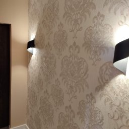 Vladyslav Vashchuk - Beżowa ściana z wzorzystą tapetą w stylu damask, oświetlona dwoma czarnymi kinkietami, widok z perspektywy korytarza z szarą podłogą i ciemnymi drzwiami po lewej stronie.