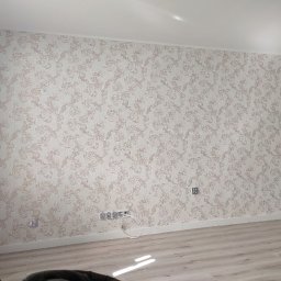 Vladyslav Vashchuk - Ściana pokryta tapetą w kremowo-złote wzory, widoczne gniazdka elektryczne i listwa przypodłogowa, jasna podłoga z paneli.