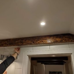 Vladyslav Vashchuk - Dekoracyjna belka z efektem postarzanego drewna zamontowana nad futryną drzwi w łazience z białymi płytkami, oświetlenie punktowe sufitowe.