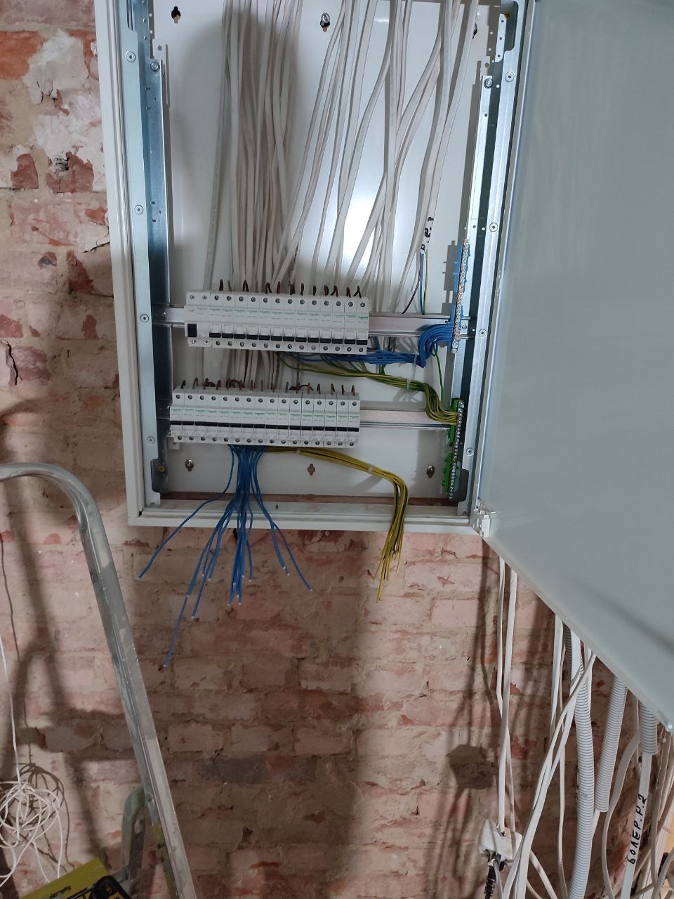 Otwarta skrzynka elektryczna z podłączonymi przewodami na tle ściany z czerwonej cegły, widoczna drabina aluminiowa.