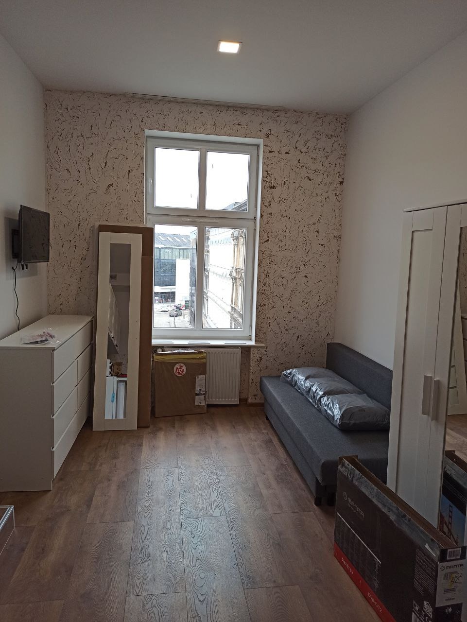 Pokój po remoncie: ściana z tapetą w odcieniach beżu i brązu, drewniana podłoga, białe meble, szara sofa z poduszkami w folii, okno z widokiem na miasto.