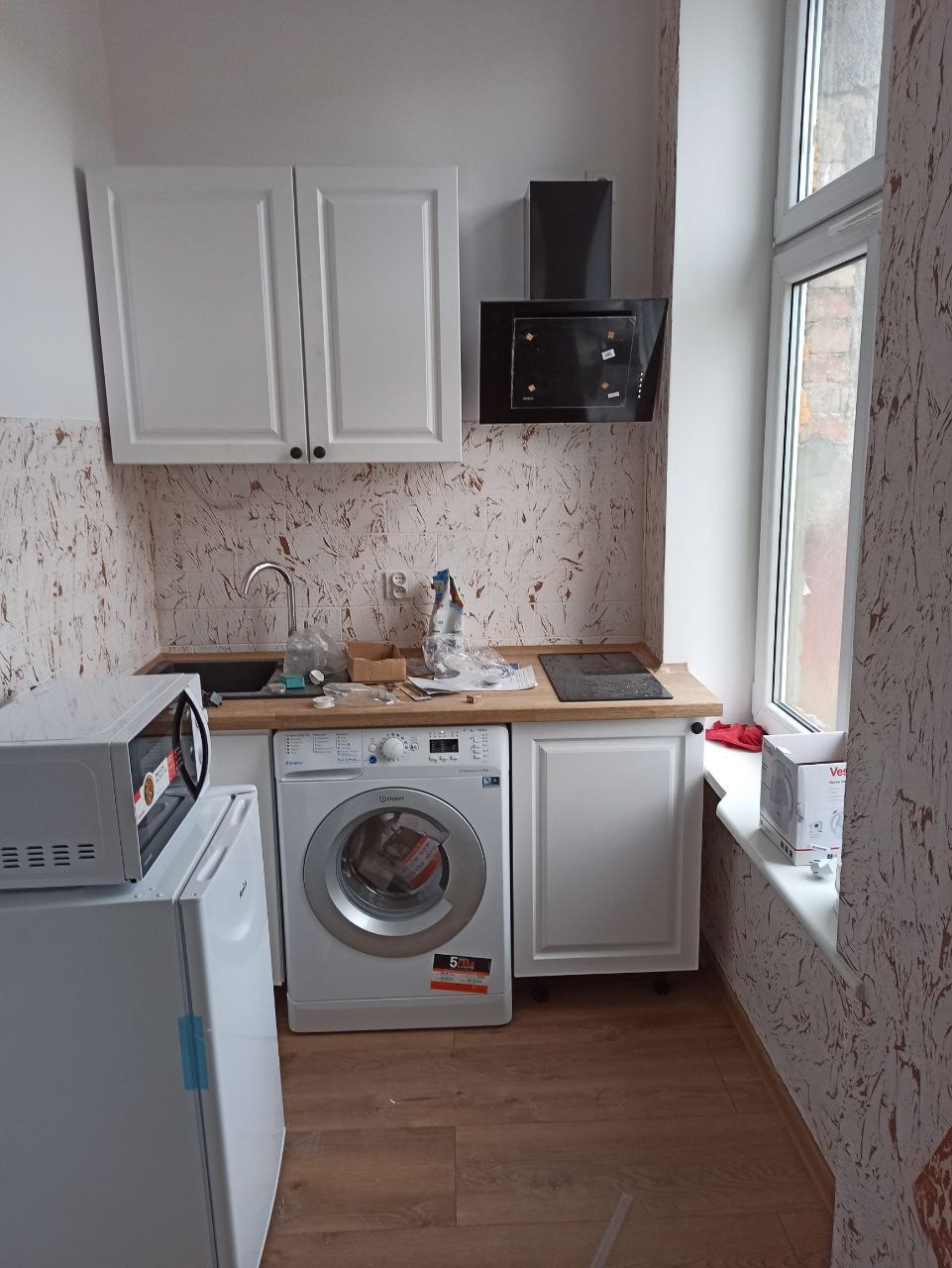 Kuchnia w trakcie prac wykończeniowych: widok na zabudowę z pralką, szafki wiszące i okap, na blacie widoczne narzędzia i akcesoria, ściany z tapetą o wzorze imitującym kamień.