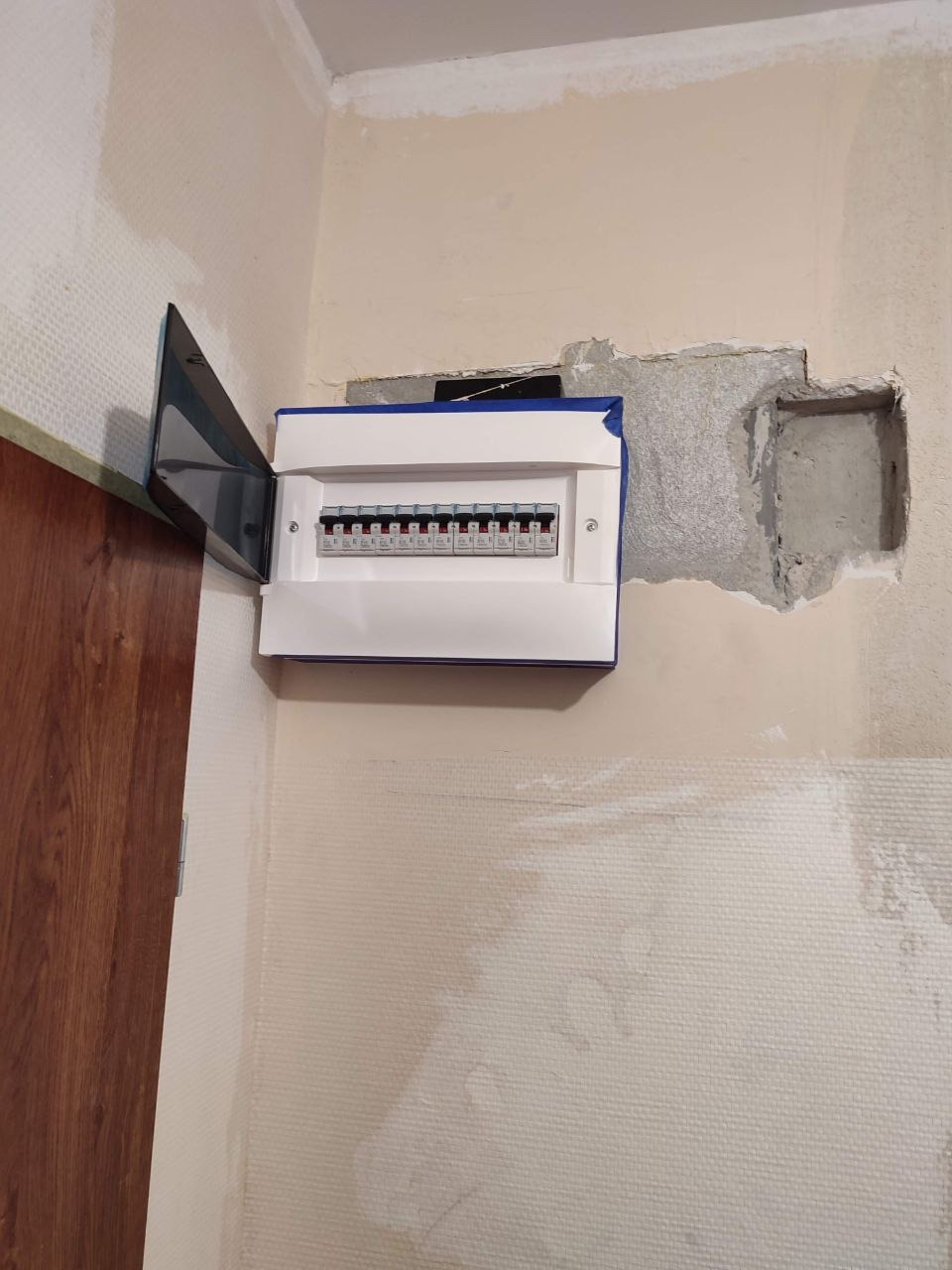 Otwarta skrzynka bezpiecznikowa zamontowana na ścianie w trakcie prac wykończeniowych, widoczne ślady gipsu i niedomalowane fragmenty tapety.