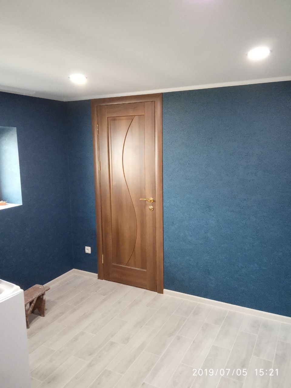 Wykończone wnętrze: ciemnoniebieskie ściany z fakturą, drewniane drzwi z ozdobnym uchwytem, jasna podłoga z paneli, widoczny fragment umywalki i stołka.