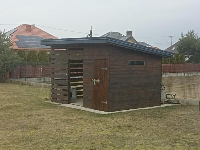 Ciemnobrązowa drewniana konstrukcja z płaskim dachem i ażurową ścianą, prawdopodobnie drewutnia lub domek narzędziowy, na trawniku w ogrodzie.