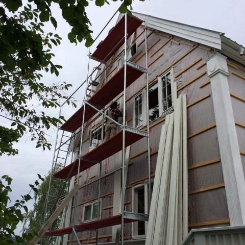 Prace wykończeniowe elewacji domu w Ostrołęce: pracownik na rusztowaniu montuje białe panele elewacyjne na ocieplonym budynku. Widoczne okna i elementy dekoracyjne.