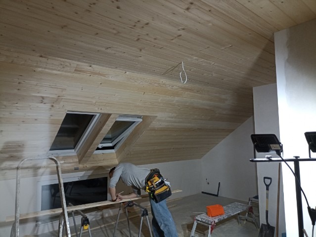 Montaż drewnianej boazerii na poddaszu z oknami dachowymi; widoczny stolarz z narzędziami, drabina oraz lampy budowlane.