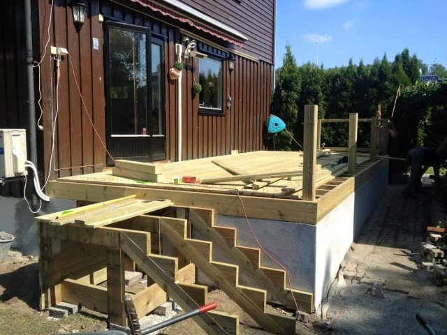 Budowa drewnianego tarasu z drewnianymi schodami przy brązowym domu z widocznymi narzędziami i poziomnicą.