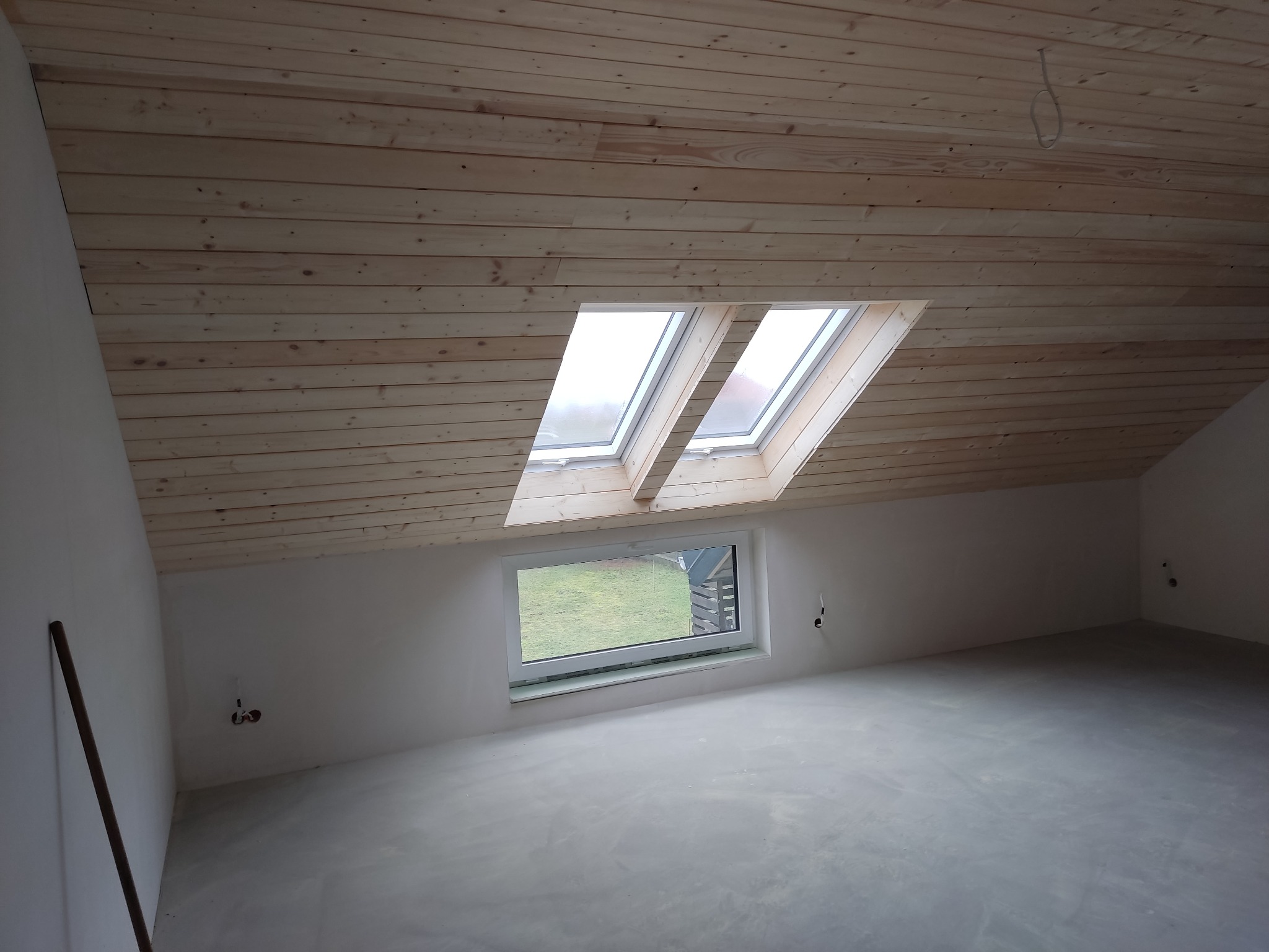 Poddasze w trakcie wykańczania: drewniana boazeria na suficie, dwa okna dachowe i jedno okno pionowe z widokiem na zieleń, surowa wylewka na podłodze.