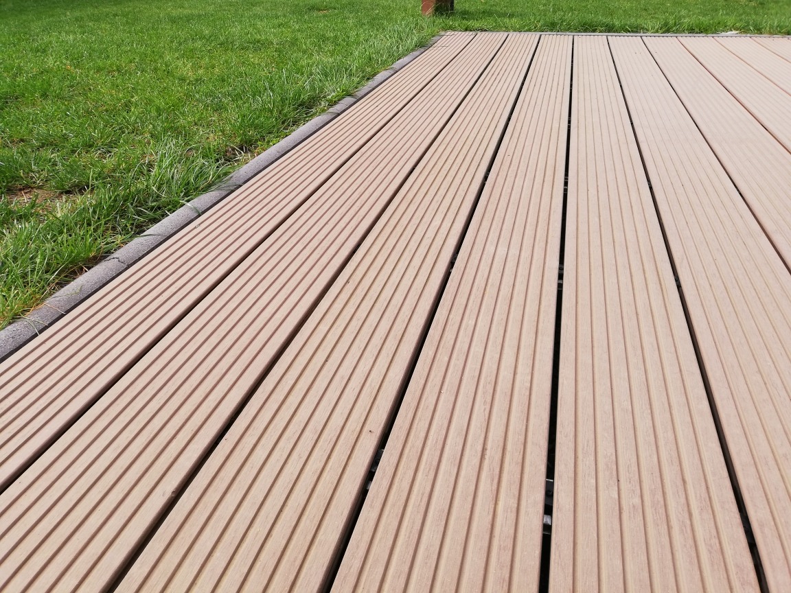 Jasna, ryflowana deska tarasowa kompozytowa ułożona na zewnątrz, obrzeże z szarej kostki, trawnik w tle.