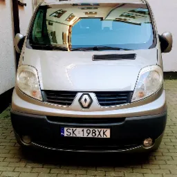 Frontowy widok srebrnego Renault Trafic z polską tablicą rejestracyjną SK 198XK, zaparkowanego na brukowanym dziedzińcu, z odbiciem budynku w przedniej szybie.