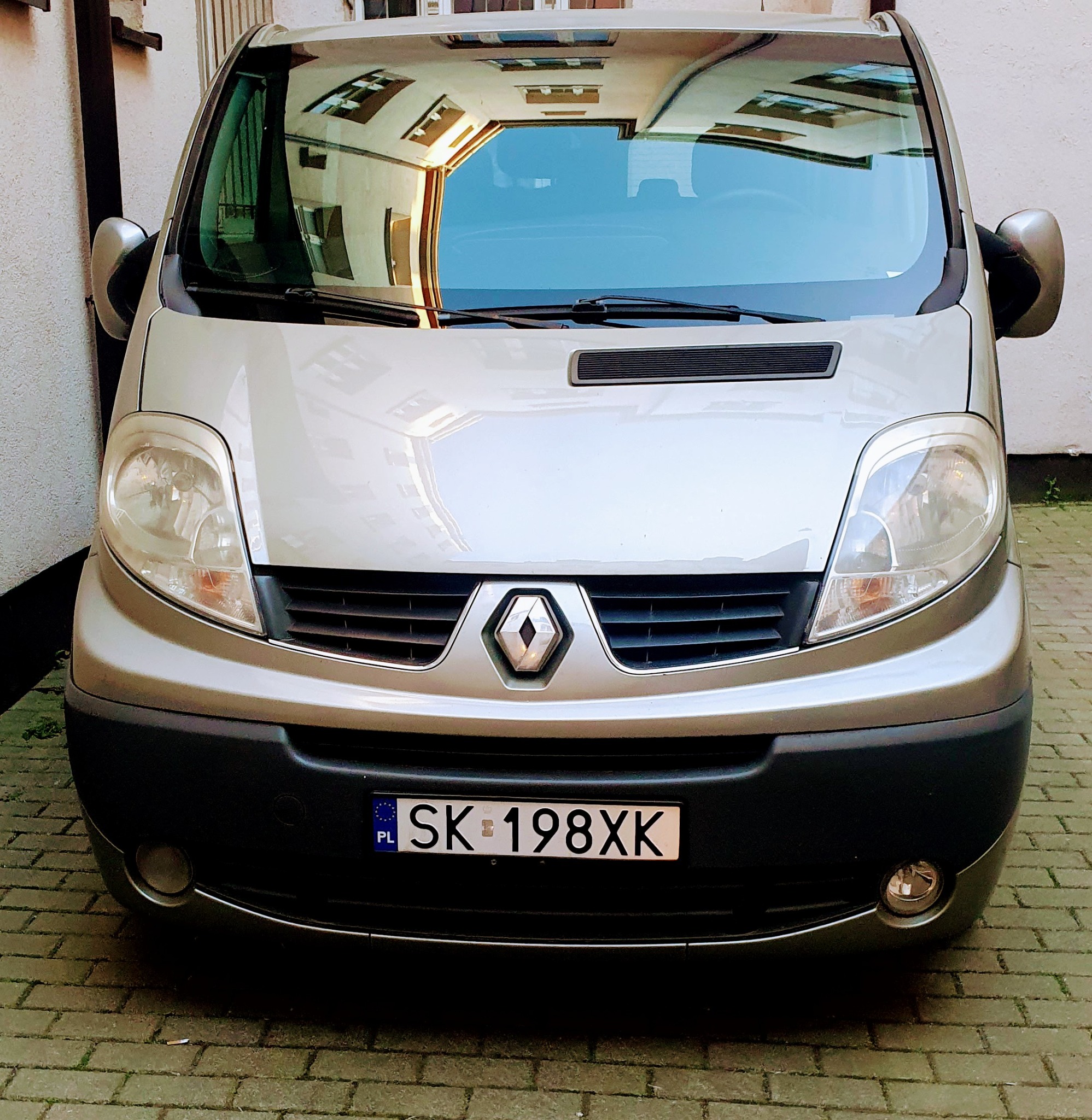 Frontowy widok srebrnego Renault Trafic z polską tablicą rejestracyjną SK 198XK, zaparkowanego na brukowanym dziedzińcu, z odbiciem budynku w przedniej szybie.