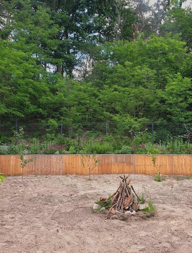 Przygotowany teren pod ogród z drewnianym ogrodzeniem i ułożonym stosem gałęzi na środku, małe drzewka posadzone wzdłuż płotu, zalesione wzgórze w tle.