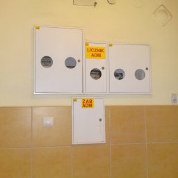 Refon Daniel Jewasiński - Zestaw białych skrzynek licznikowych z żółtymi etykietami 'Licznik ADM' i 'ZAB ADM' zamontowanych na ścianie nad brązowymi płytkami, widoczne liczniki przez okrągłe otwory.