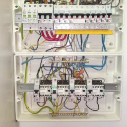 Otwarta, dwuczęściowa, biała skrzynka elektryczna z zamontowanymi bezpiecznikami, różnokolorowymi przewodami i listwami zaciskowymi, widok z góry.